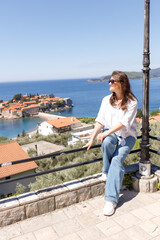 Young woman in casual clothes sitting on the railing of the observation deck overlooking the sea and the island in the background, summer vacation, travel and tourism concept, space for text