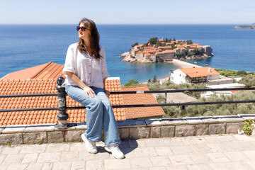 Young woman in casual clothes sitting on the railing of the observation deck overlooking the sea and the island in the background, summer vacation, travel and tourism concept, space for text