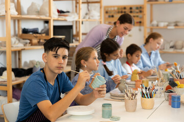 Obraz premium Teenager paints a clay mug with brush at a workshop with a teacher. Boy at a ceramics master class works at a table in an art studio