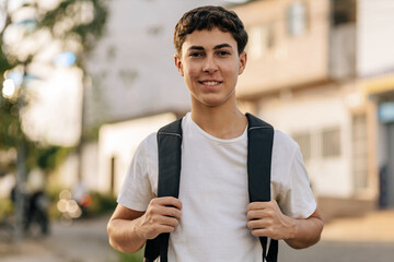 Teenage student standing outdoors in an urban neighborhood wearing a backpack and smiling at the camera. Concept of youth, education, daily routine, independence, mobility and modern lifestyle
