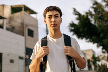 Teenage student standing outdoors in an urban neighborhood wearing a backpack and smiling at the camera. Concept of youth, education, daily routine, independence, mobility and modern lifestyle