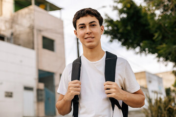 Teenage student standing outdoors in an urban neighborhood wearing a backpack and smiling at the camera. Concept of youth, education, daily routine, independence, mobility and modern lifestyle