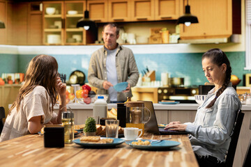 Busy mother ignoring her daughter at breakfast and focusing on her work, typing information on laptop and looking preoccupied. Distant working mom with a career and tween girl.