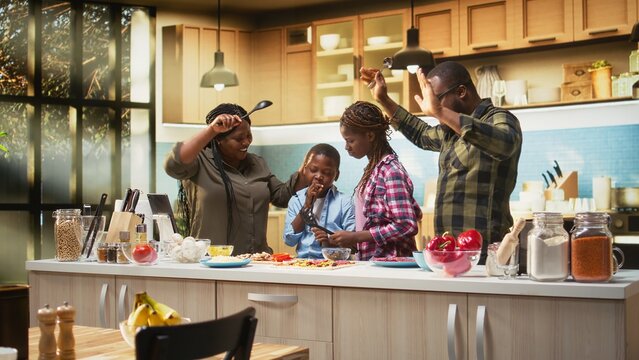 Black parents and kids having fun singing and dancing in the kitchen, cooking homemade pizza and acting goofy for the bonding weekend activity. Happy playful family at home. Camera A.