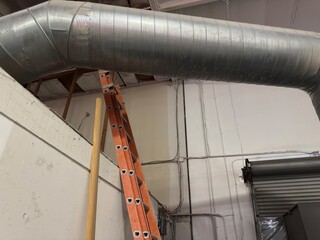 Naklejka premium Industrial Interior Detail: View of large HVAC ductwork, an orange safety ladder, and the top of a commercial garage door.