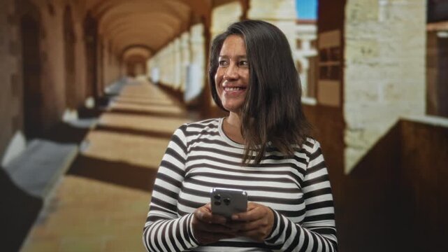 Woman holding smartphone with hands visible, smiling and looking at screen in a sandstone building corridor; contentment connection.