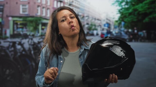 Middle aged woman holding motorcycle helmet, puckering lips and raising hand on a street; confidence adventure.