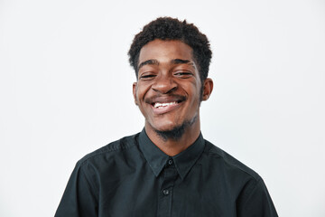 Obraz premium Young black man with dark skin smiling and wearing black shirt on white background. Portrait of happy African ethnicity male with short curly hair and closed eyes.