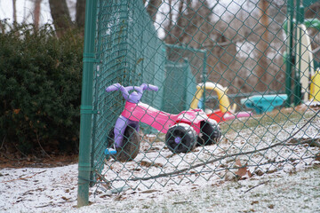 Children's pink plastic tricycle toy abandoned in a fenced yard area during the winter season with fresh snow on the ground.