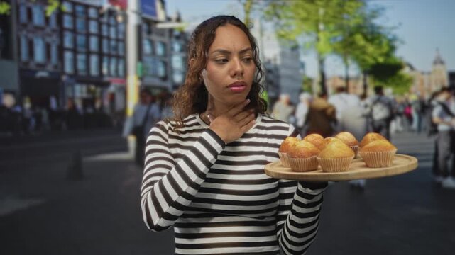 Young african american woman holding wooden tray of muffins, hand on throat in crowded city street; concern food safety.