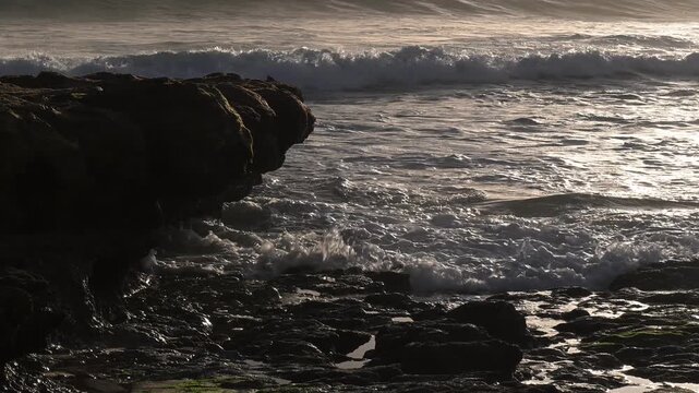 Waves crash against rocks at sunset along the coast during a cloudy evening
