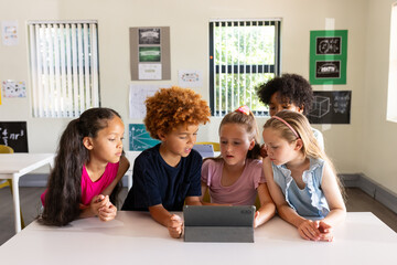 Diverse schoolchildren gathering around tablet on white table in classroom, studying with posters