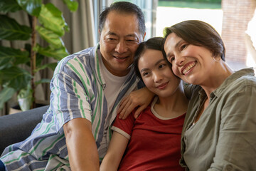 Mature parents with adult daughter sitting on dark couch by potted plant in living room, smiling