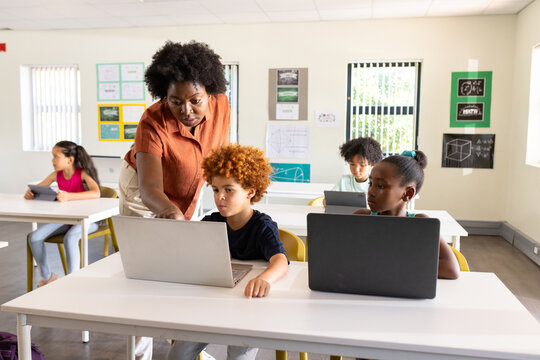 African teacher with school-age students leaning over desk assisting boy using laptop in classroom