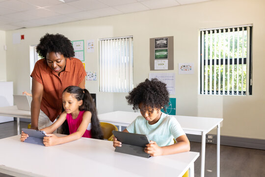 African American teacher leaning over girl and boy kids using tablets at white desks with posters