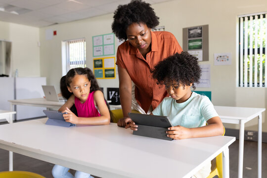 African American woman teacher leaning over table supervising two children using tablets in class