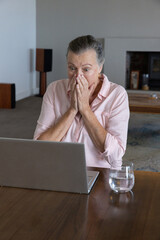 Naklejka premium Senior woman sitting at living-room table in pink shirt using open laptop, covering mouth, glass