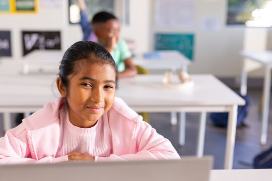 African American preteen classmates sitting at desks in classroom, girl using laptop in pink hoodie