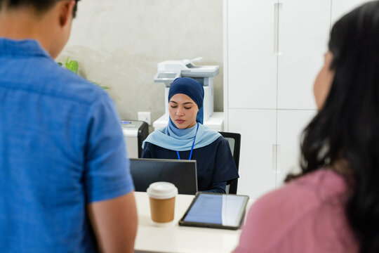 Diverse receptionist wearing blue headscarf talking with clients at reception counter on laptop