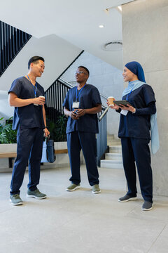 Diverse coworkers chatting wearing navy scrubs and lanyards in atrium, holding tablet and coffee