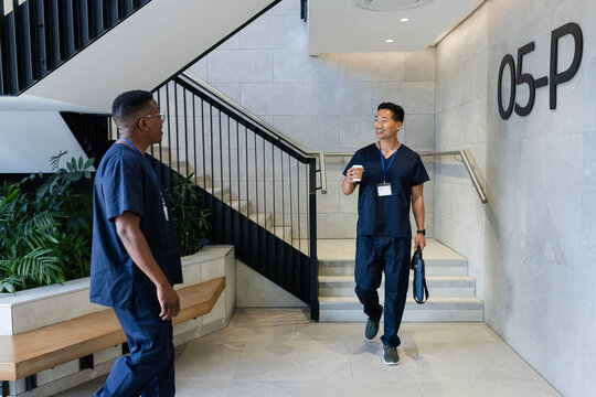 Diverse male coworkers in navy scrubs, blue IDs, chatting on stairs holding paper cup and bag