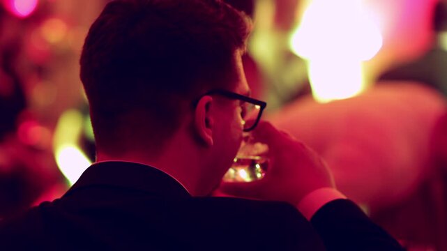 Guest in a Suit and Glasses Taking a Sip at the Table with Dancing Party Crowd in the Background