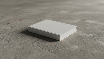 A rectangular, blank book rests on a rough, textured concrete surface, casting a subtle shadow