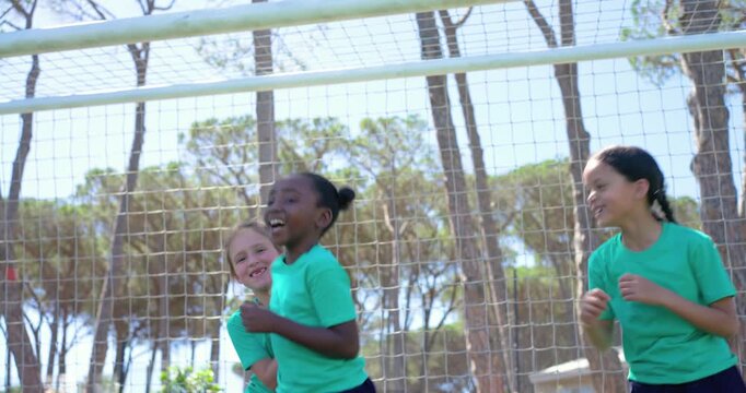 Diverse female child team in green shirts celebrating scoring goal, bouncing at soccer net