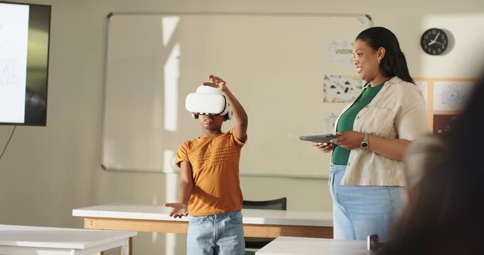 Teacher using tablet while African youth donning VR headset, reaching virtual objects in classroom