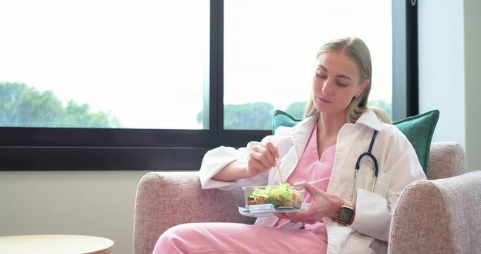 Adult female nurse in scrubs on break in lounge, stirring salad from clear container, recharging