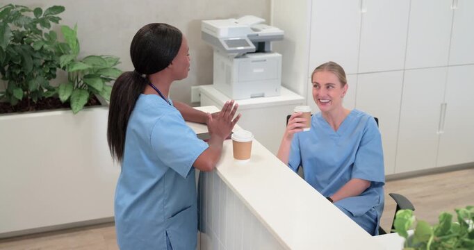 Diverse female nurses in scrubs chatting at clinic, standing nurse placing cup gesturing on break