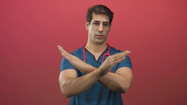 Man doctor wearing scrubs and stethoscope crossing forearms and hands in x stop gesture in studio red backdrop; safety warning.