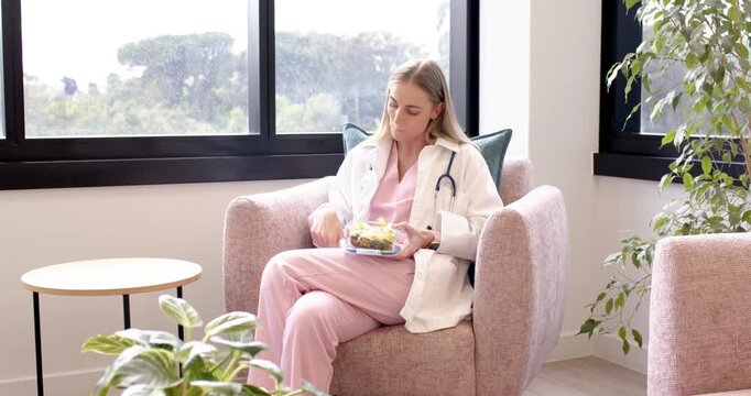 Female nurse in scrubs, stethoscope eating salad, pausing for alert, reading phone in clinic room