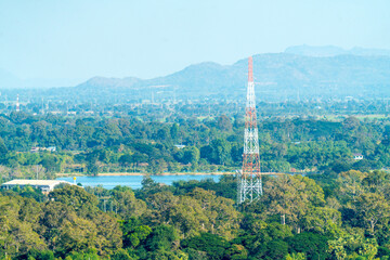 Large telecommunication tower standing tall in a lush green forest landscape with mountains and a lake in the background. Remote area signal coverage and rural connectivity infrastructure.