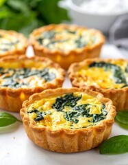 Mini spinach quiches on white surface, bright yellow filling, flaky crust, with fresh green leaves