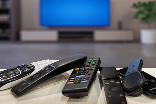 A group of generic tv remote controls on a table with a widescreen tv in the background of a living room.