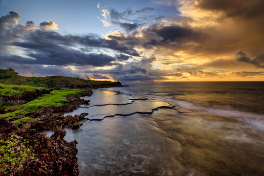 Sunrise at Inarajan Pools on Guam as the sun peaks out of orange-colored clouds in this long exposure blurring the waves