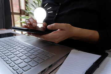 A woman is working on a laptop and holding a mobile phone. The image shows a light bulb icon with a luminous effect. The scene is bright and dynamic.