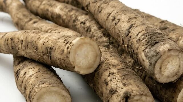 A close-up shot captures a fresh pile of unwashed root vegetables, prominently displaying their natural, earthy brown skin against a clean white background. The long, slender roots are intricately tex