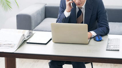 A male business person talking on a smartphone while looking at a PC in the office