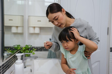 stubborn child girl refusing to brush her teeth in the bathroom. mother is trying to encourage her unhappy daughter during the morning routine, while child looks grumpy and resistant.