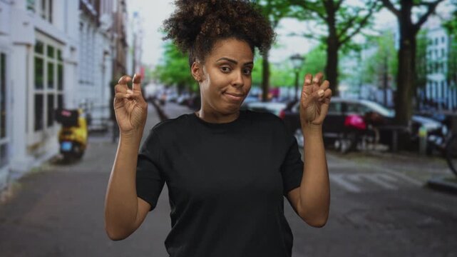 Woman making air quotes with fingers on a city street, hands raised, wearing black tee and curly hair, face expressed with mild smirk; sarcasm commentary.