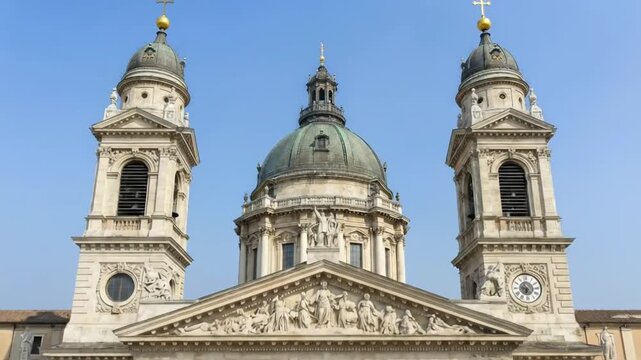 A majestic historical building with a grand central dome and two ornate towers stands against a clear blue sky. Its intricate stone facade, adorned with classical columns, detailed carvings, and statu