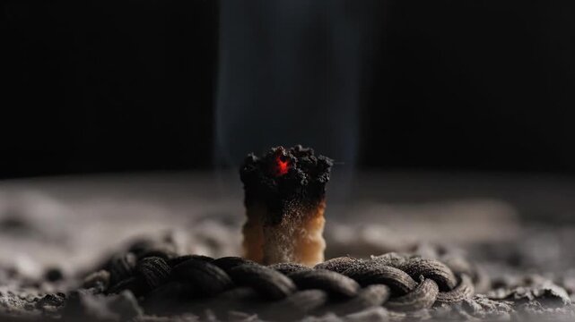 Close up Extreme Macro Shot Of A Burning Candle Wick With Orange Flame And Wisps Of Smoke Ascending Against A Dark Background and Ash Scattered Around The Base