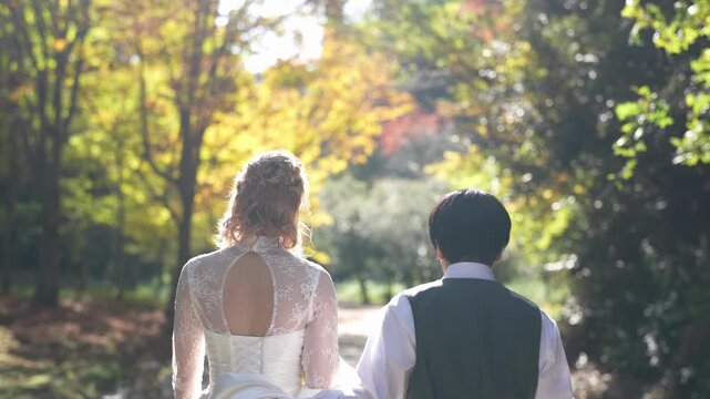 A bride and groom are walking together in a forest. The bride is wearing a white dress and the groom is wearing a vest. Scene is romantic and peaceful