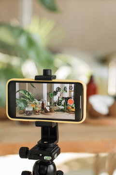 A smartphone mounted on a tripod is recording a table filled with fresh fruits, jars of ingredients, and plants, suggesting content creation for food blogging or vlogging