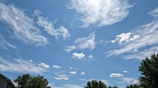 A beautiful, clear blue sky is filled with an array of dynamic white clouds, ranging from delicate, wispy cirrus formations to scattered, fluffy cumulus puffs. The bright sun occasionally creates a ge