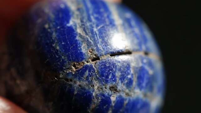 Close Up Macro Shot Of A Textured Lapis Lazuli Sphere With Golden Pyrite Inclusions Held Gently By A Human Finger Against A Dark Background With Natural Lighting And Sharp Focus