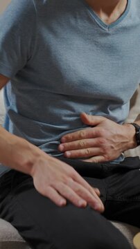 Man sitting on sofa in living room holding stomach and suffering from abdominal pain or cramps. Person suffering from health issues, digestion problems or food poisoning at home