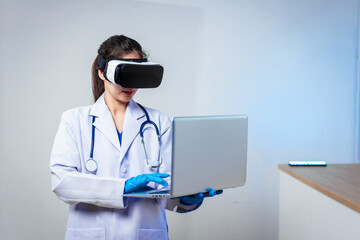 An Asian female doctor wearing a virtual reality headset uses a laptop in  modern clinic,demonstrating innovative medical technology,digital healthcare training,telemedicine solutions,future-focused
